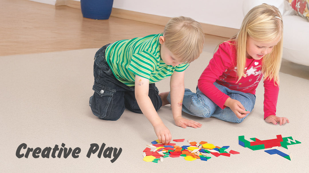 Two children engaged in creative play on the floor, using colorful geometric shapes to build patterns and figures. Text overlay reads 'Creative Play'.